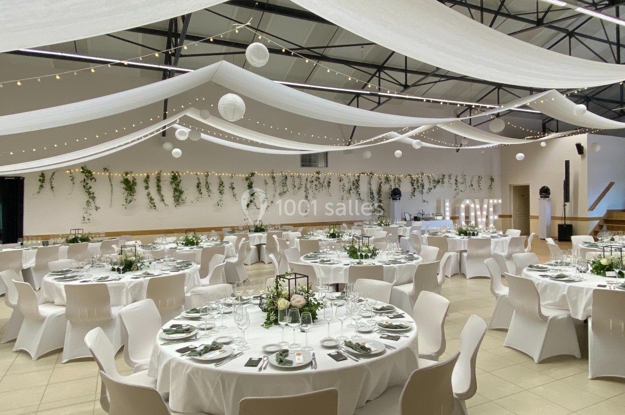 Salle de réception décorée avec des tables rondes, nappes blanches, guirlandes lumineuses et suspensions végétales.