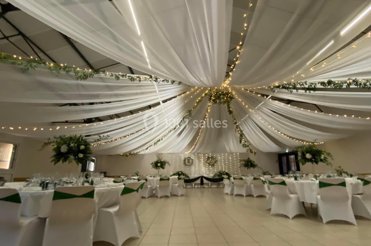 Salle de réception décorée avec des drapés blancs suspendus, guirlandes lumineuses et tables élégamment dressées.
