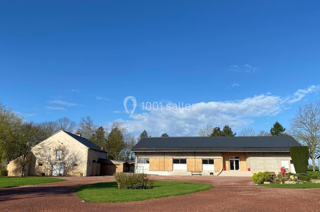 Bâtiments ruraux avec façades claires, entourés de pelouse, arbres et ciel bleu dégagé.