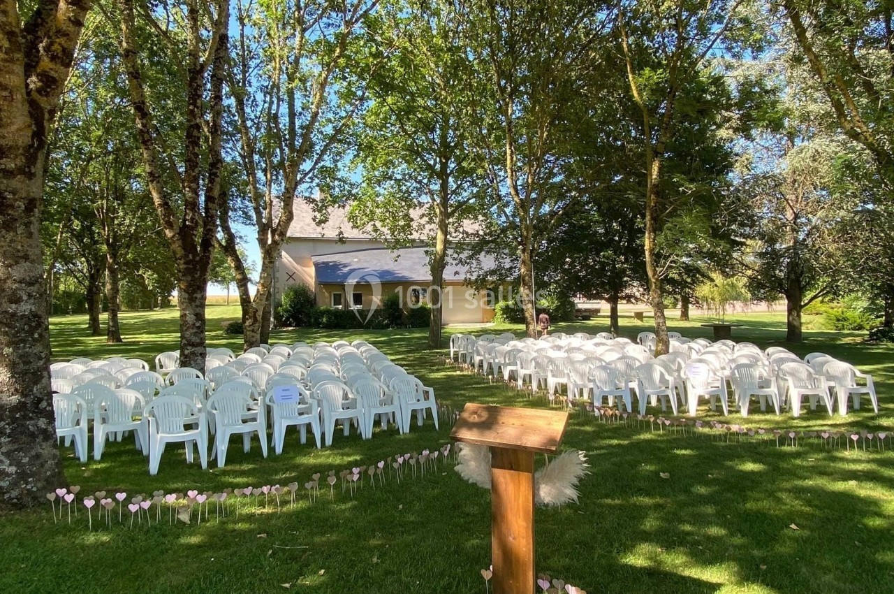 Chaises blanches disposées en rangées sur une pelouse ombragée, face à un pupitre en bois, près d'un bâtiment.