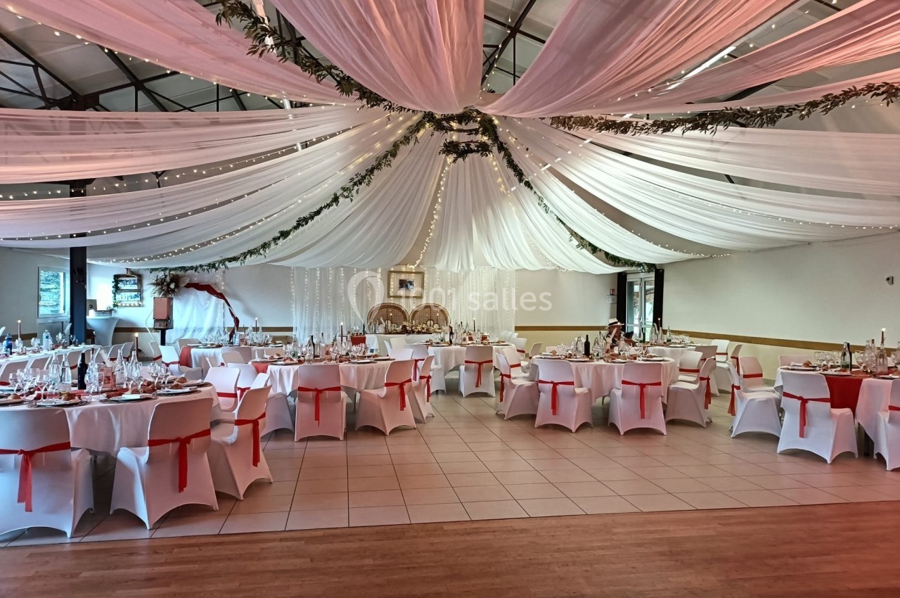 Salle de réception décorée avec des drapés blancs au plafond, tables rondes dressées et chaises ornées de nœuds rouges.