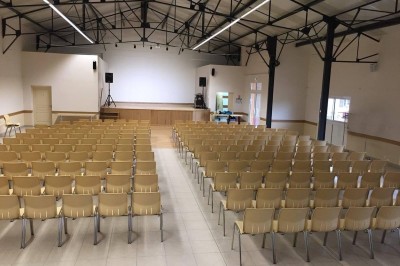 Salle avec des rangées de chaises en plastique beige face à une scène surélevée dans un espace lumineux.