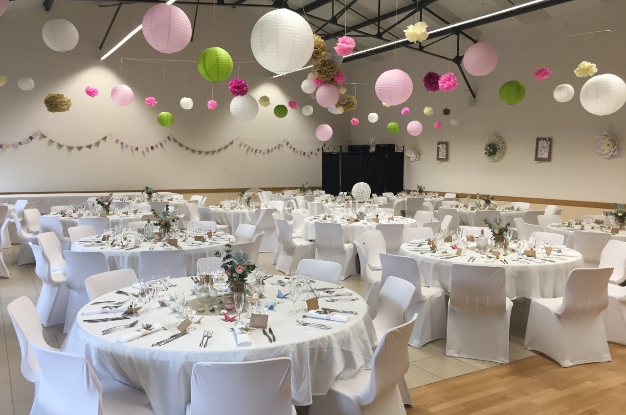 Salle de réception décorée avec des tables rondes dressées, des lanternes et des fleurs suspendues au plafond.