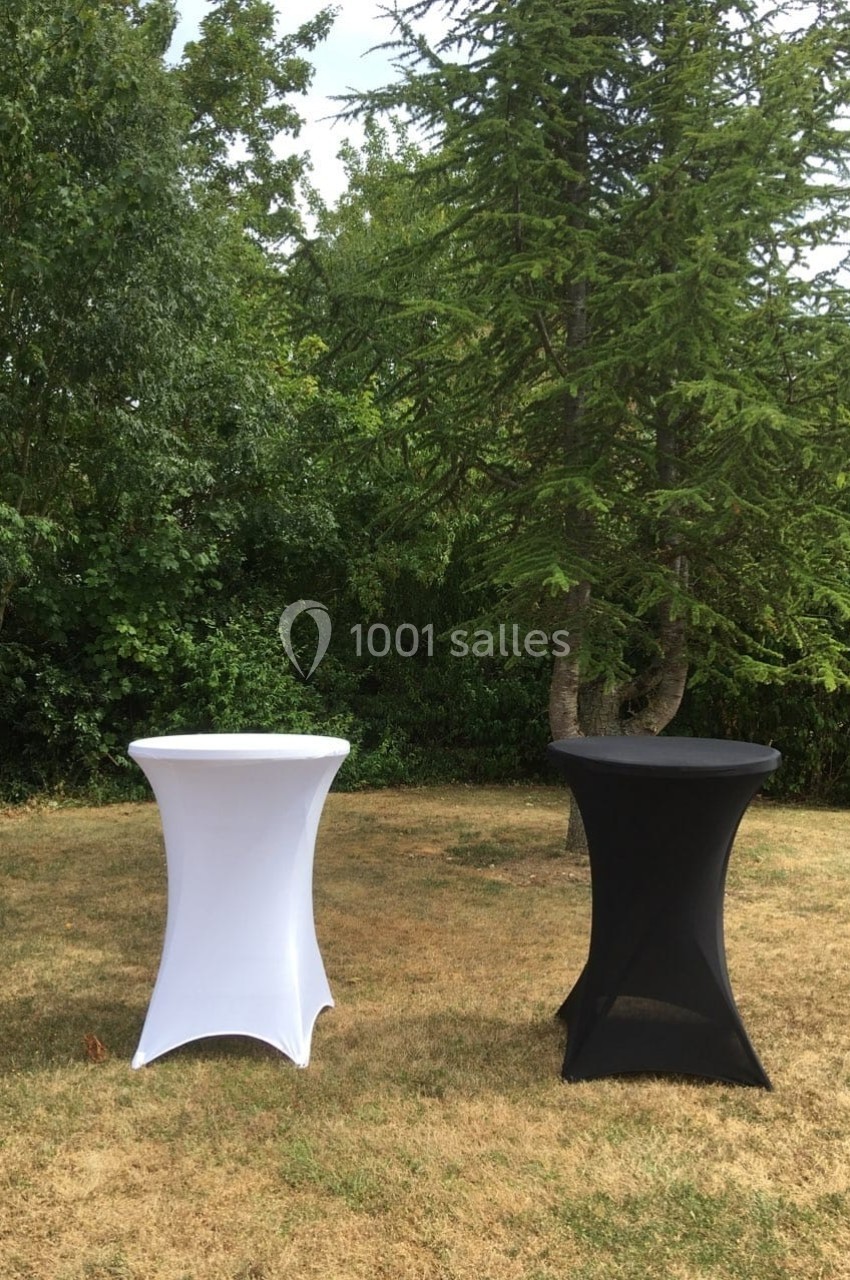 Deux tables hautes recouvertes de housses blanches et noires sont disposées sur une pelouse devant des arbres.