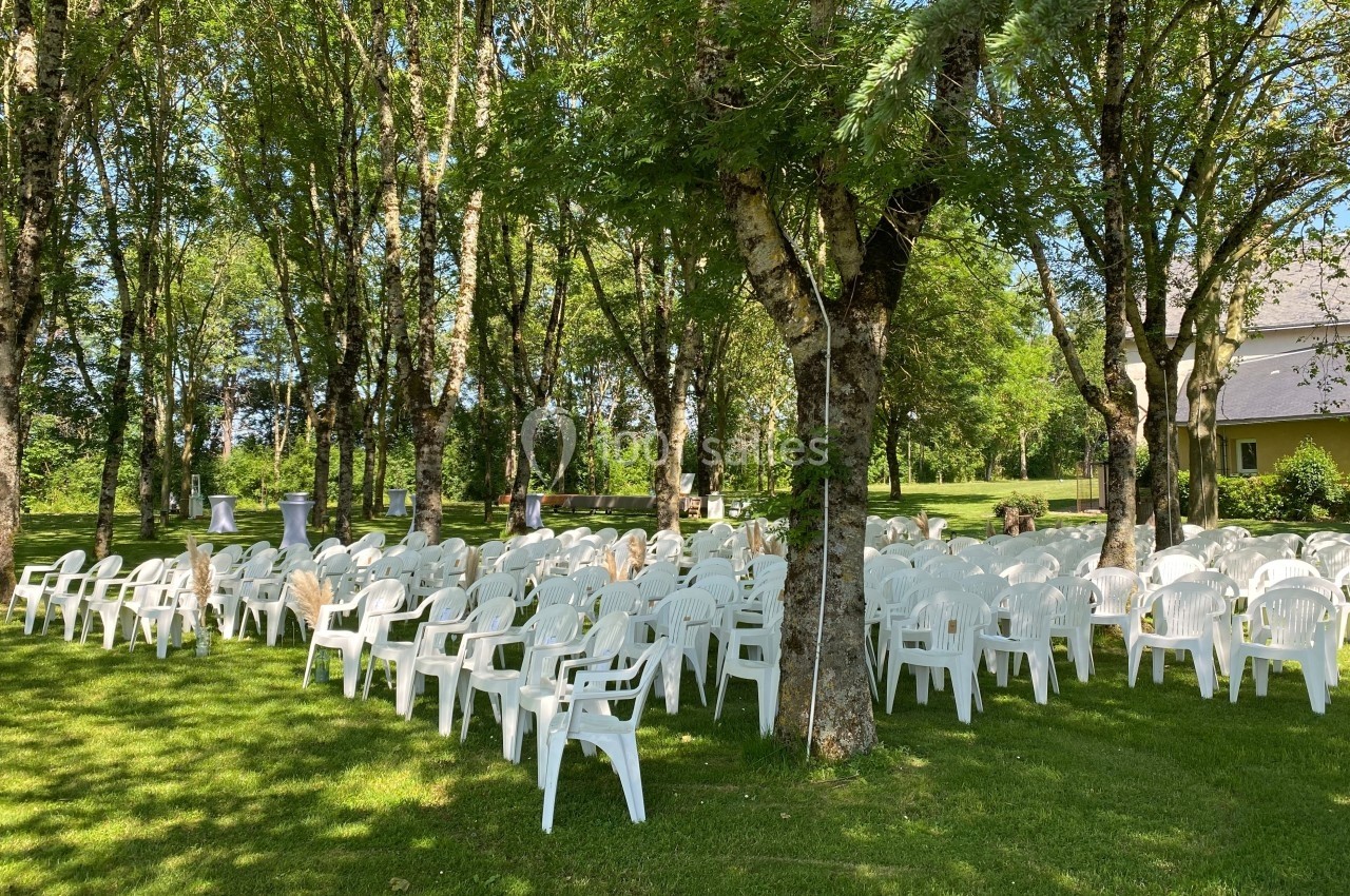 Chaises blanches disposées en rangées sur une pelouse ombragée par des arbres, préparées pour un événement en extérieur.