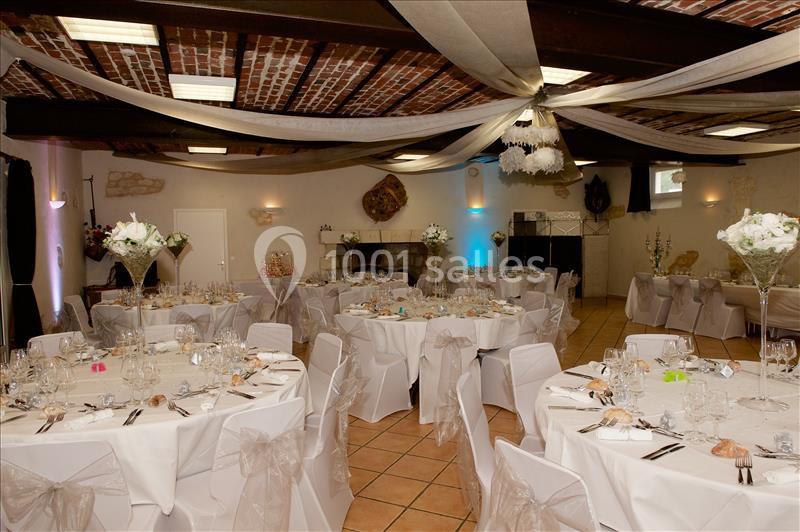 Salle de réception décorée avec des tables rondes, nappes blanches, chaises ornées de nœuds et plafonds en poutres…
