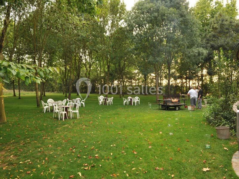 Des chaises blanches disposées sur une pelouse entourée d'arbres, avec un barbecue allumé et des personnes à proximité.