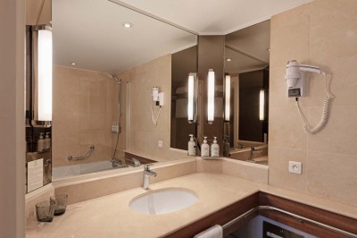 Chambre d'hôtel lumineuse avec lit double, bureau, fauteuil, grandes fenêtres et vue sur un balcon.