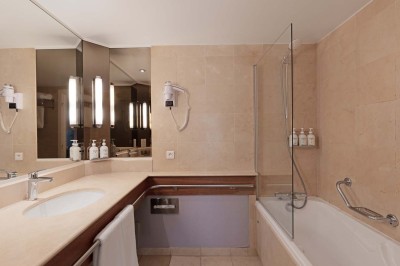 Chambre d'hôtel lumineuse avec lit double, bureau, fauteuil, grandes fenêtres et vue sur un balcon.