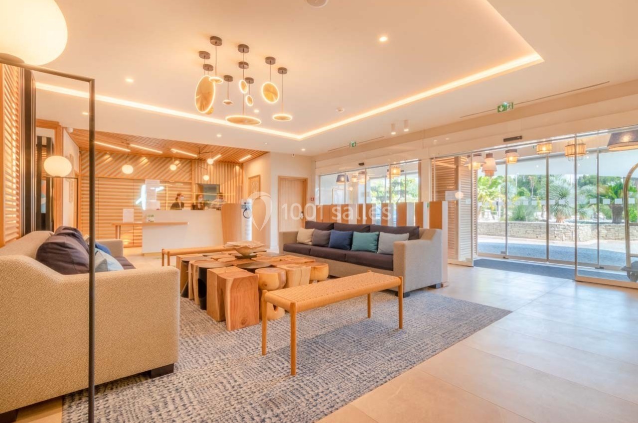 Espace d'accueil lumineux avec mobilier en bois, canapés, tapis bleu et grandes baies vitrées donnant sur un jardin.