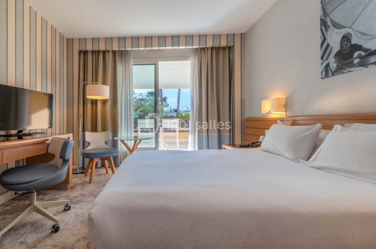 Chambre d'hôtel lumineuse avec lit double, bureau, fauteuil, télévision et balcon donnant sur un espace extérieur verdoyant.