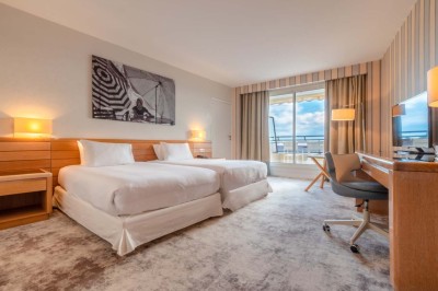 Chambre d'hôtel lumineuse avec lit double, bureau, fauteuil, grandes fenêtres et vue sur un balcon.