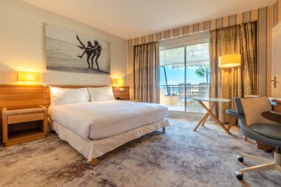 Chambre d'hôtel lumineuse avec lit double, bureau, fauteuil, grandes fenêtres et vue sur un balcon.