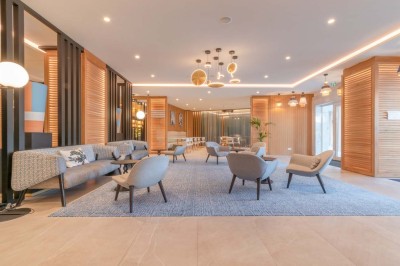 Chambre d'hôtel lumineuse avec lit double, bureau, fauteuil, grandes fenêtres et vue sur un balcon.
