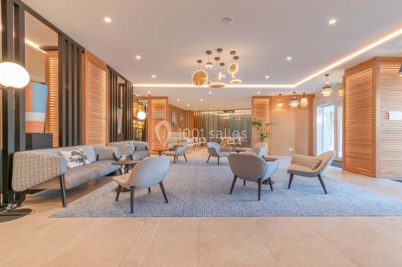 Espace lounge lumineux avec fauteuils, canapés, tapis bleu et éclairage moderne, entouré de panneaux en bois.