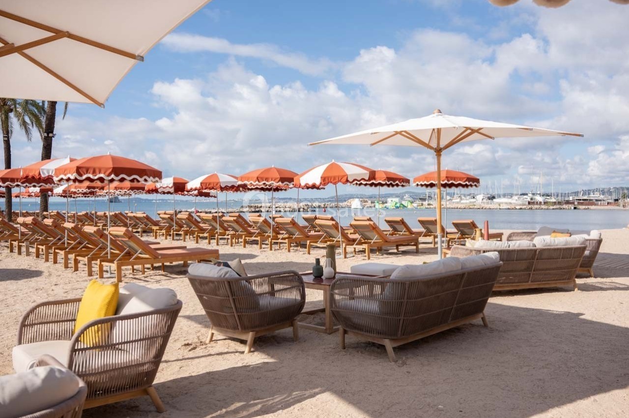 Chaises longues et parasols orange sur une plage de sable avec vue sur la mer et un port en arrière-plan.