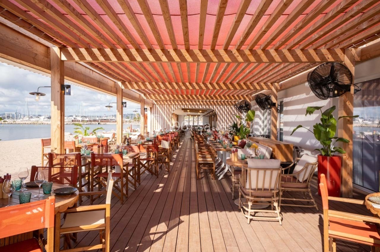 Terrasse en bois d'un restaurant en bord de plage, avec tables et chaises disposées sous une pergola.