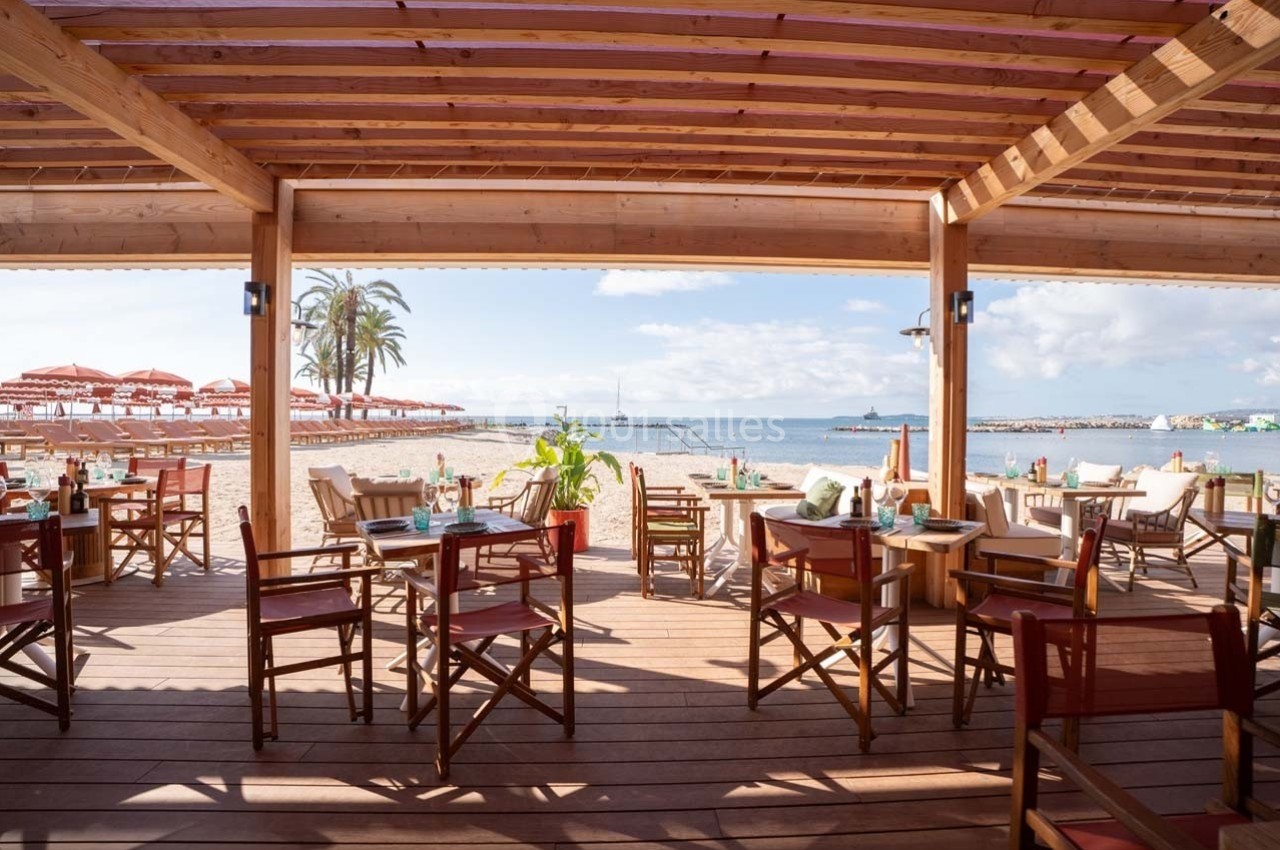 Terrasse en bois d'un restaurant en bord de mer avec vue sur la plage, des tables dressées et des parasols rouges.