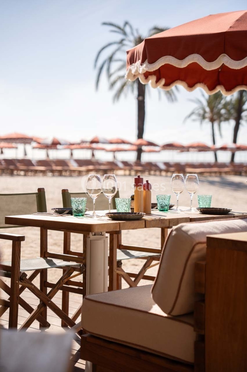 Table dressée avec verres et condiments sous un parasol rouge, en bord de plage avec palmiers et transats en arrière-plan.