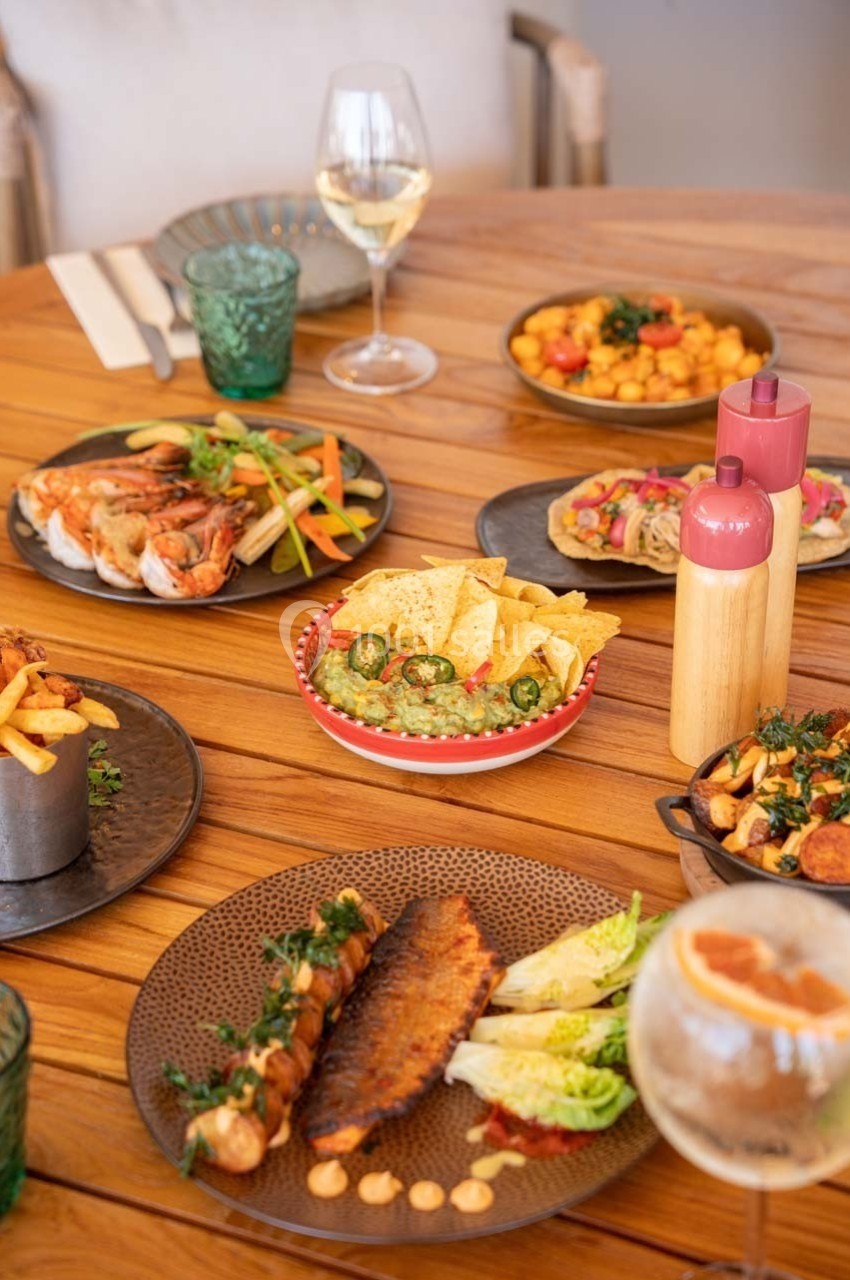 Table en bois garnie de plats variés, dont des fruits de mer, tacos, guacamole, frites et verres de boisson.