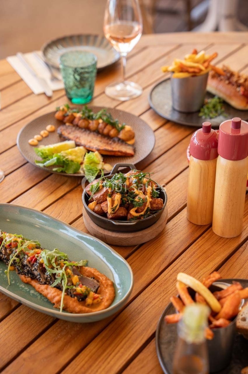 Table en bois dressée avec des plats variés, incluant viandes, légumes, frites et condiments, accompagnés de boissons.