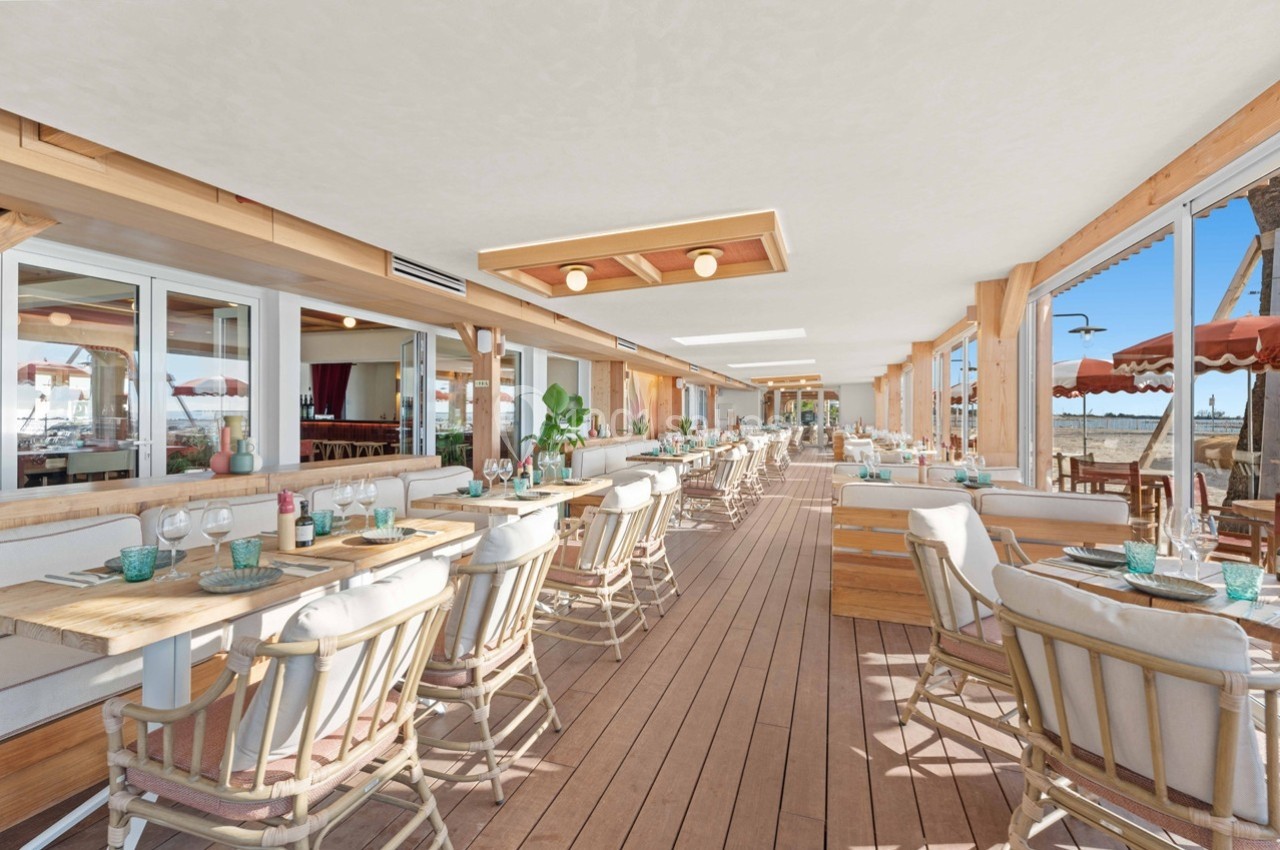 Salle de restaurant lumineuse avec mobilier en bois, grandes baies vitrées et vue sur l'extérieur ensoleillé.