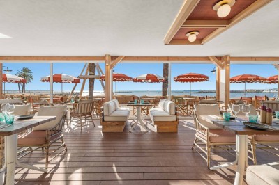Balcon avec table et chaises blanches, vue sur des palmiers, la mer et un ciel bleu, sous un toit en panneaux blancs.