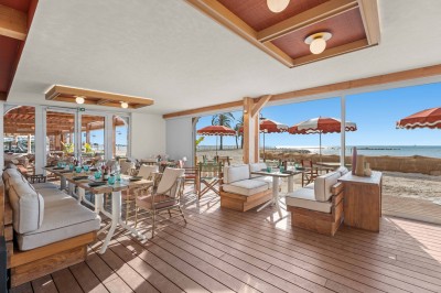 Balcon avec table et chaises blanches, vue sur des palmiers, la mer et un ciel bleu, sous un toit en panneaux blancs.