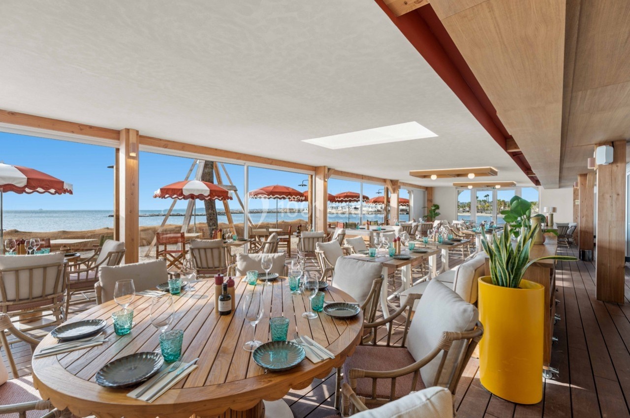 Salle de restaurant lumineuse avec vue sur la plage, tables en bois dressées et parasols rouges à l'extérieur.
