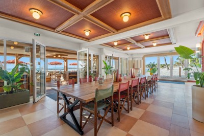 Balcon avec table et chaises blanches, vue sur des palmiers, la mer et un ciel bleu, sous un toit en panneaux blancs.