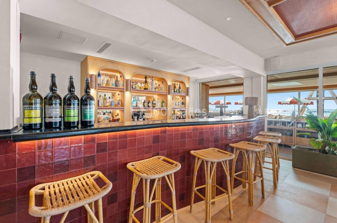Bar avec comptoir en carrelage rouge, tabourets en rotin, étagères de bouteilles et vue sur une terrasse ensoleillée.