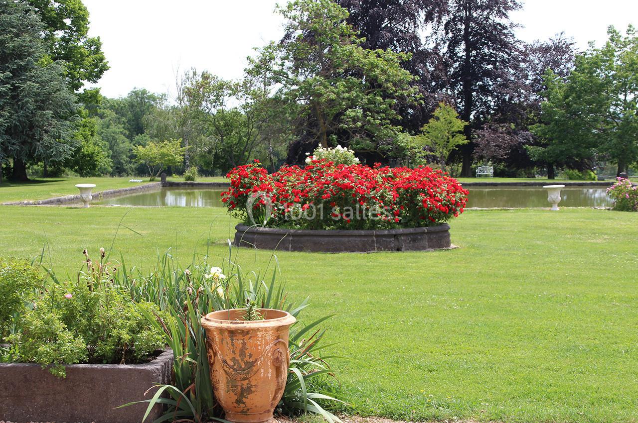Jardin verdoyant avec un parterre de fleurs rouges, un pot en terre cuite au premier plan et un bassin en arrière-plan.
