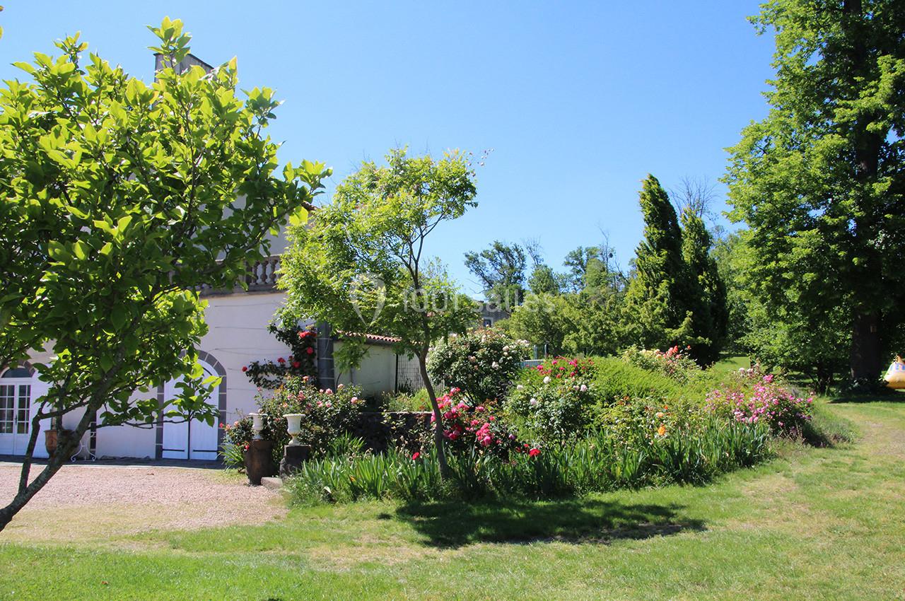 Jardin fleuri avec pelouse, arbustes et arbres, bordant une maison blanche sous un ciel bleu clair.