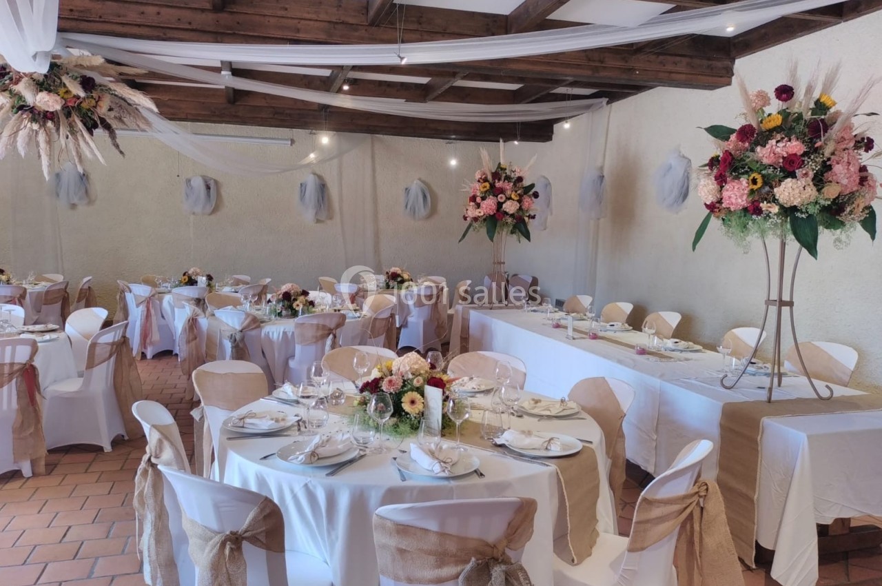 Salle de réception décorée avec des tables rondes, nappes blanches, chaises ornées de rubans et arrangements floraux.