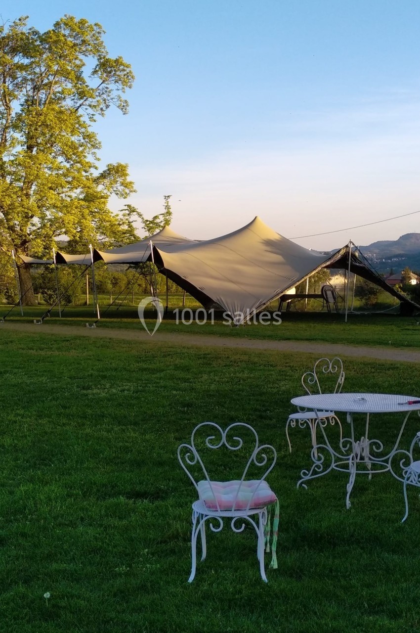 Tente beige installée sur une pelouse verdoyante avec des chaises et une table en fer forgé au premier plan.