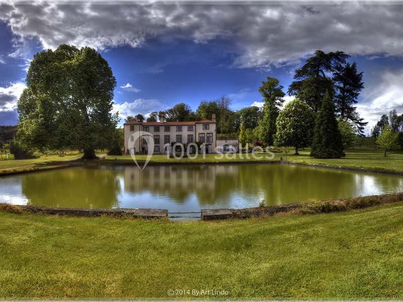 Manoir entouré d'arbres, reflété dans un étang calme sous un ciel partiellement nuageux.