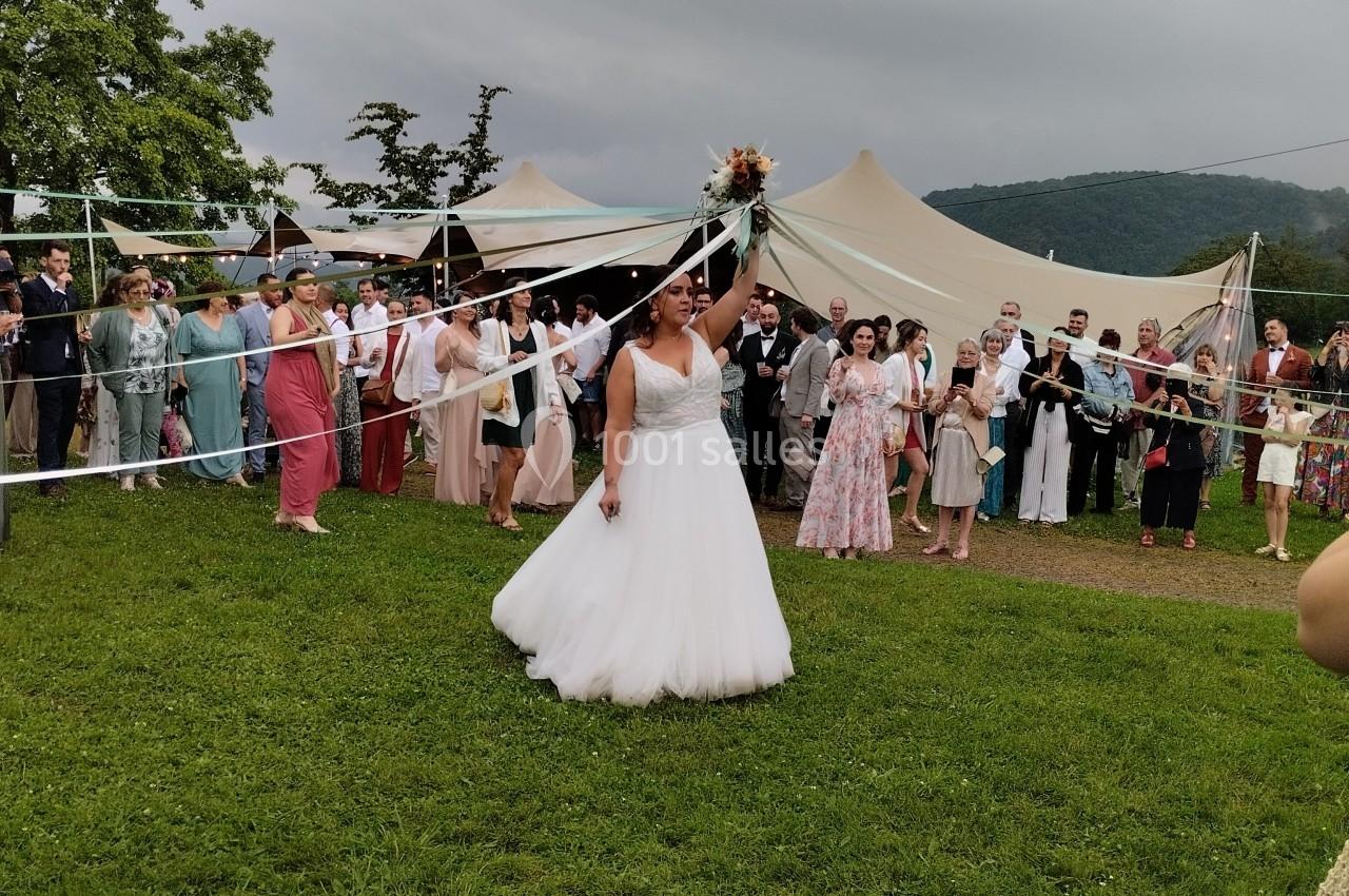 Une mariée en robe blanche lance son bouquet devant un groupe de convives dans un cadre extérieur verdoyant.