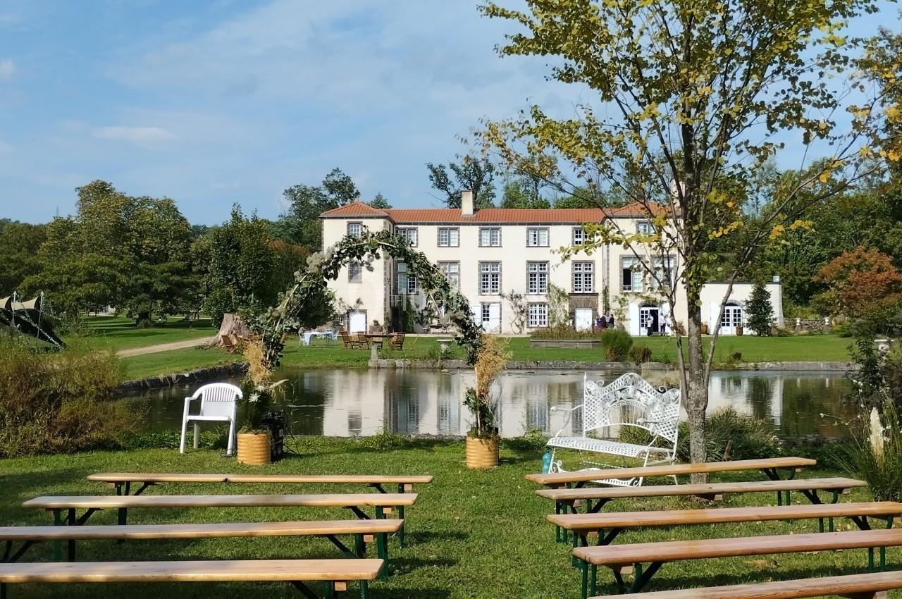 Bancs en bois disposés en rangées sur une pelouse face à un étang et une grande maison blanche entourée de verdure.