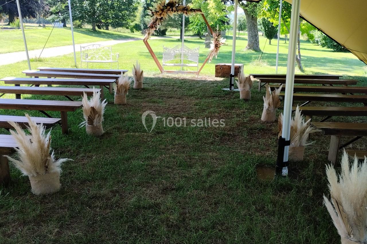 Allée centrale d'une cérémonie en plein air sous une tente, décorée de bancs en bois et de bouquets de pampas.