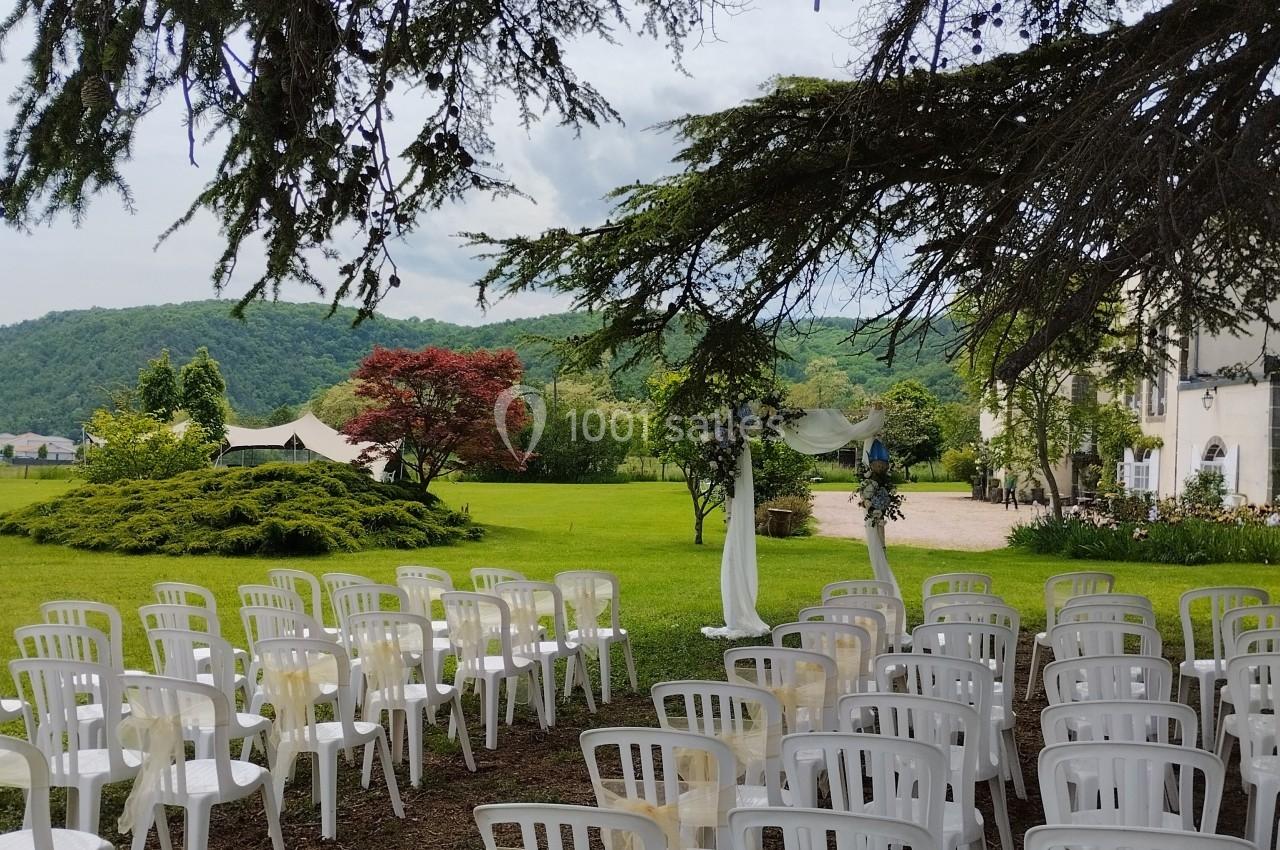 Chaises blanches disposées en rangées sous un arbre, face à un espace vert avec une tente et des collines en arrière-plan.