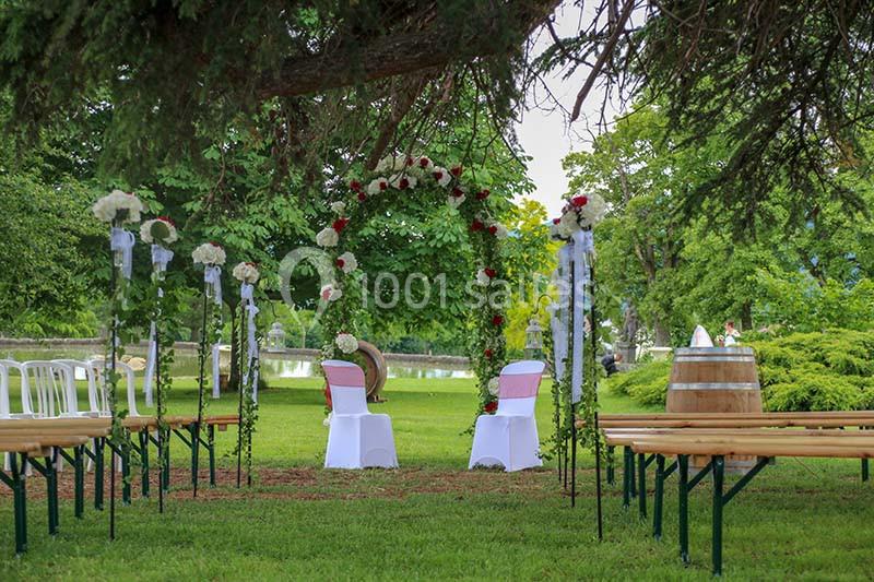 Chaises décorées sous une arche fleurie dans un jardin verdoyant, entourées de bancs en bois pour une cérémonie en plein air.