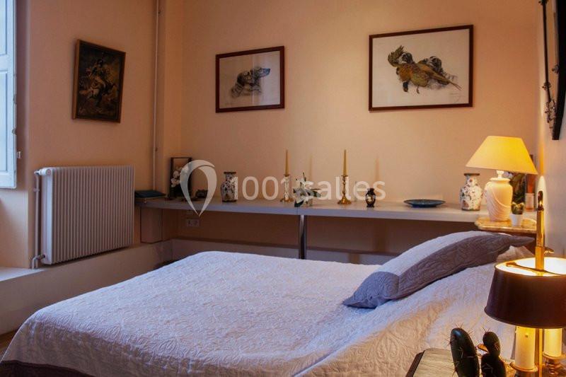 Chambre avec un lit double, murs beige, décorations murales, lampe allumée et étagères ornées de vases et chandeliers.