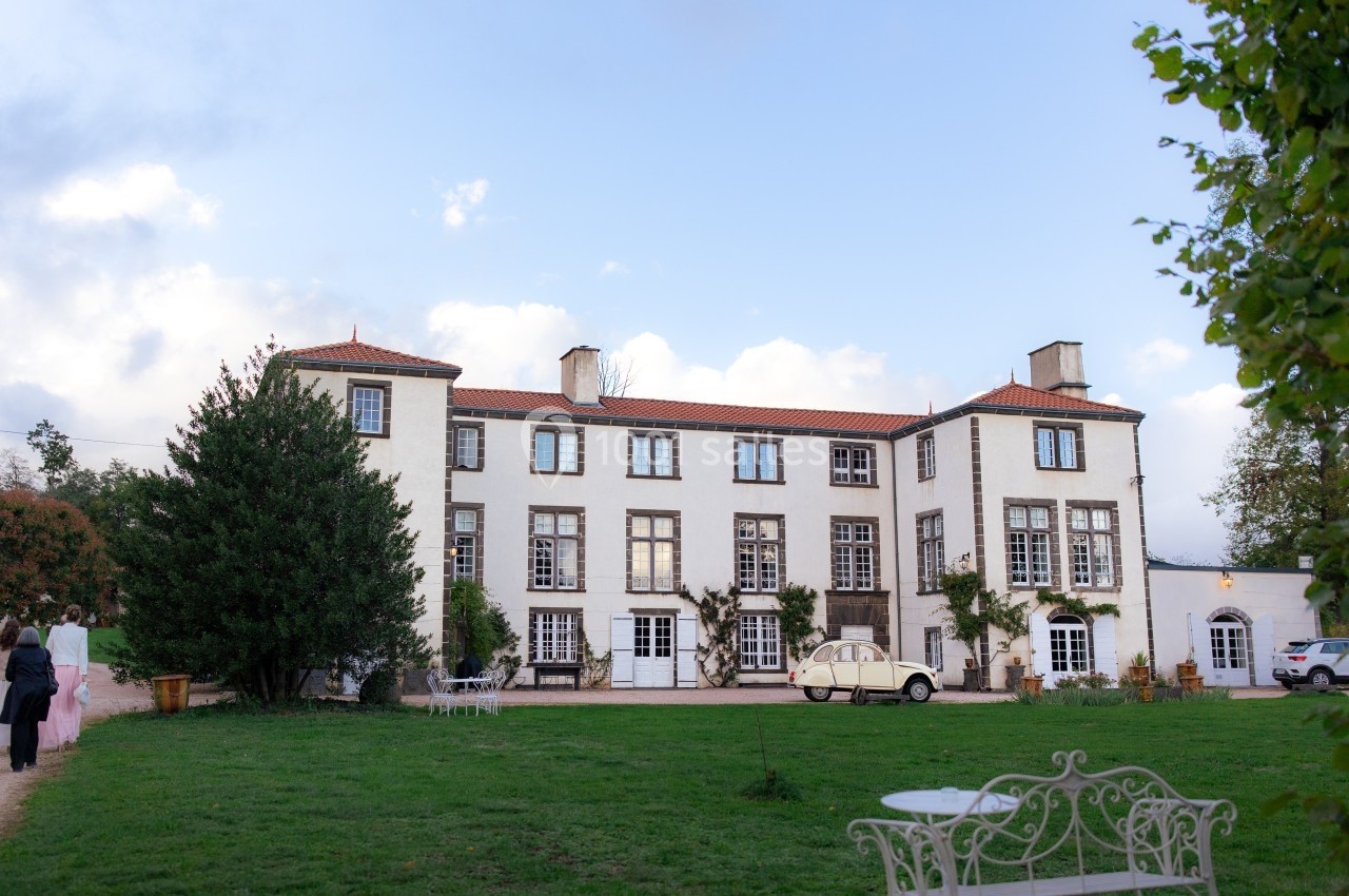 Façade d'un grand bâtiment blanc à trois étages avec un jardin verdoyant et une voiture ancienne stationnée devant.