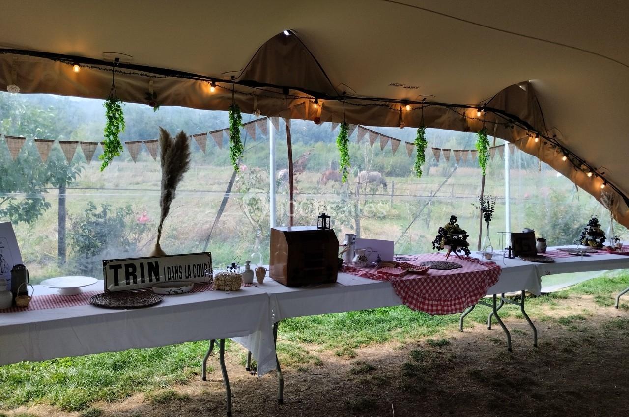 Tables décorées avec nappes rouges et blanches sous une tente, avec vue sur un champ et des guirlandes suspendues.