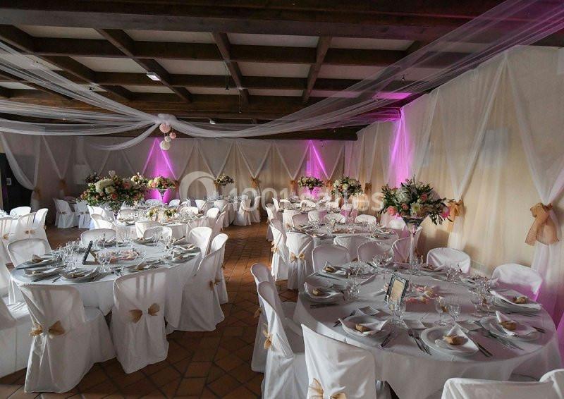 Salle de réception décorée pour un mariage avec tables rondes, nappes blanches, fleurs et éclairage rose.