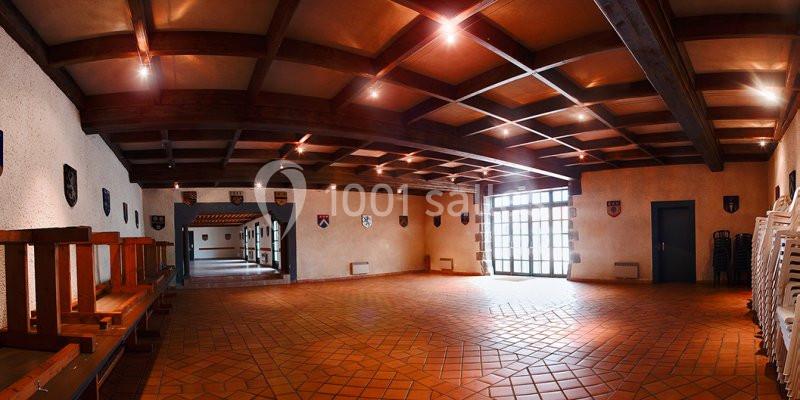 Salle spacieuse avec carrelage au sol, poutres apparentes au plafond, murs décorés de blasons et chaises empilées.