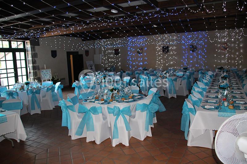 Salle de réception décorée avec des tables rondes, nappes blanches et nœuds bleus, éclairée par des guirlandes lumineuses.
