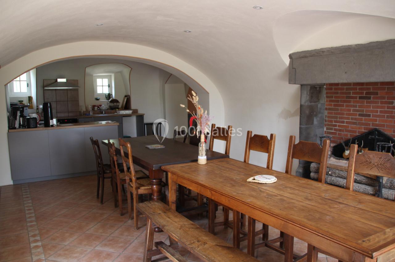 Salle à manger avec une grande table en bois, chaises assorties, cheminée en pierre et cuisine ouverte en arrière-plan.