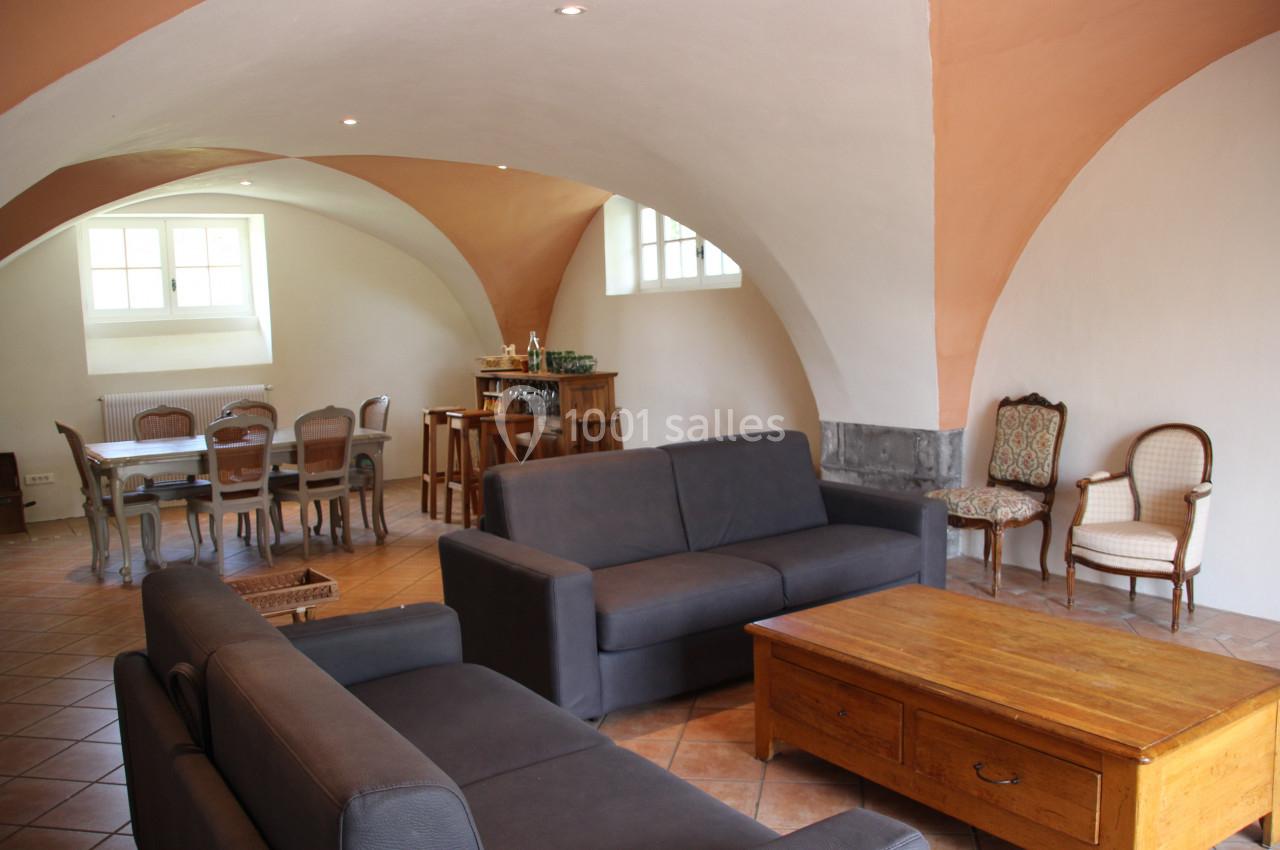 Salle voûtée avec canapés gris, table basse en bois, table à manger et chaises anciennes sur un sol en carrelage.