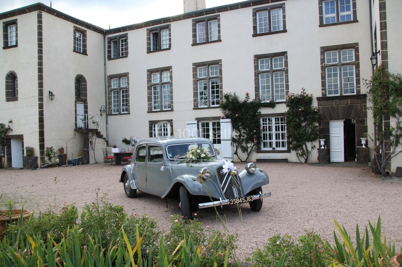 Voiture ancienne grise décorée de fleurs stationnée devant une grande bâtisse aux façades blanches et fenêtres à croisillons.